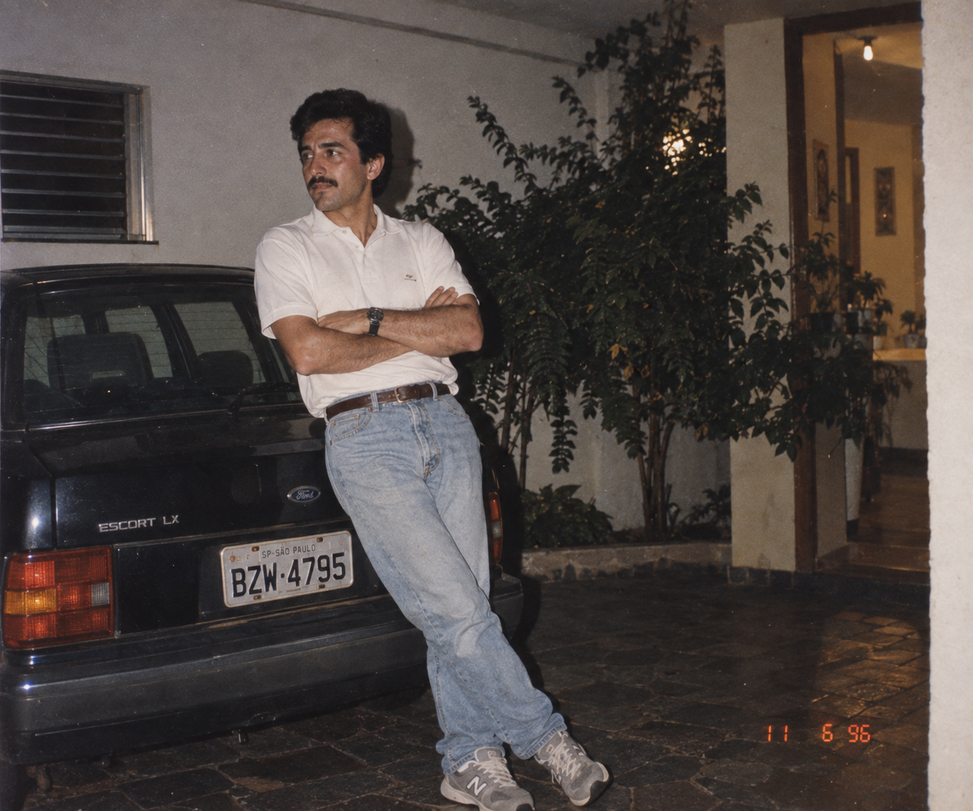 Homem com polo branca, jeans claro e New Balance encostado em um Ford Escort em 1996